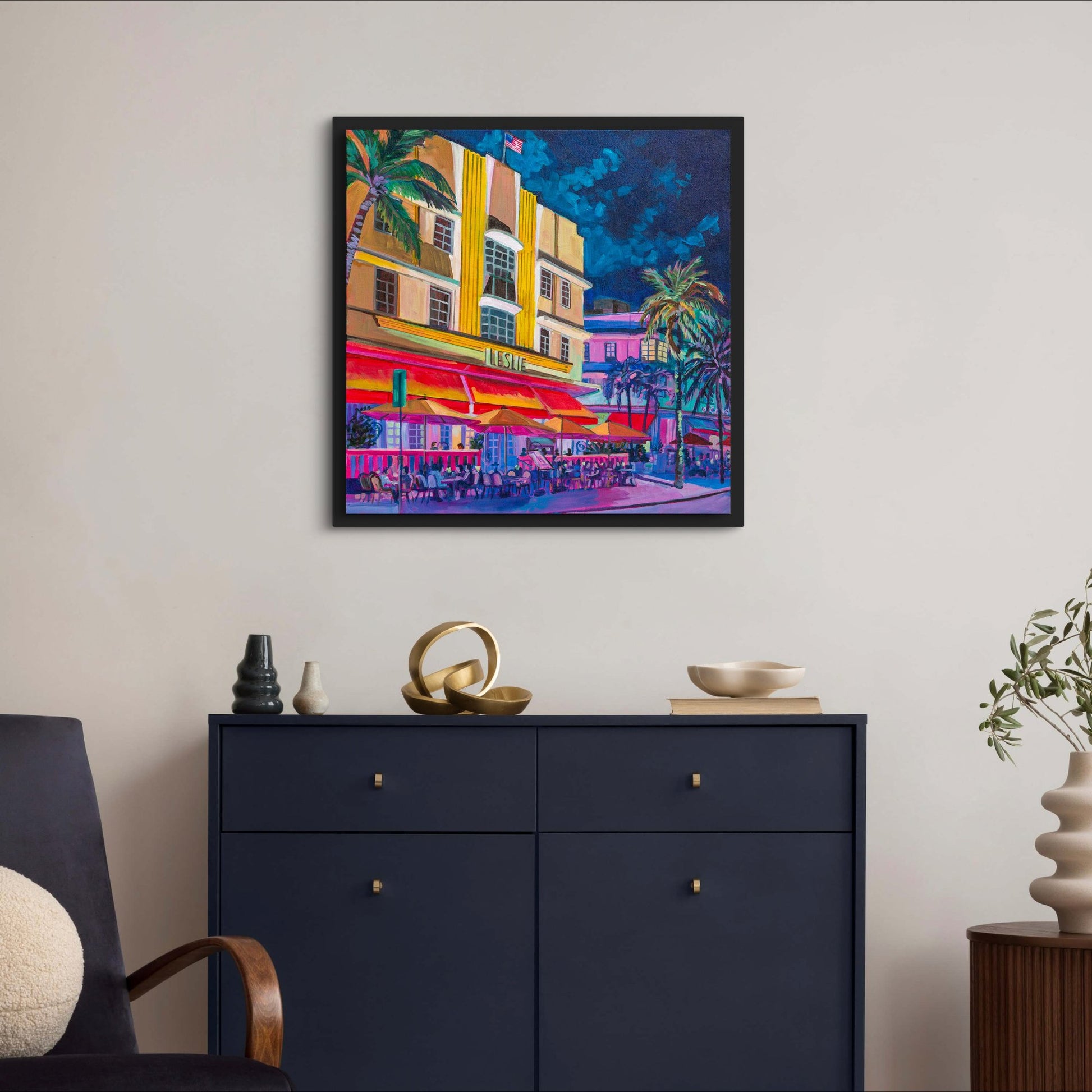 Architectural painting of iconic art deco hotels with dramatic lighting at night hangs in sleek living room area with navy credenza and modern vases
