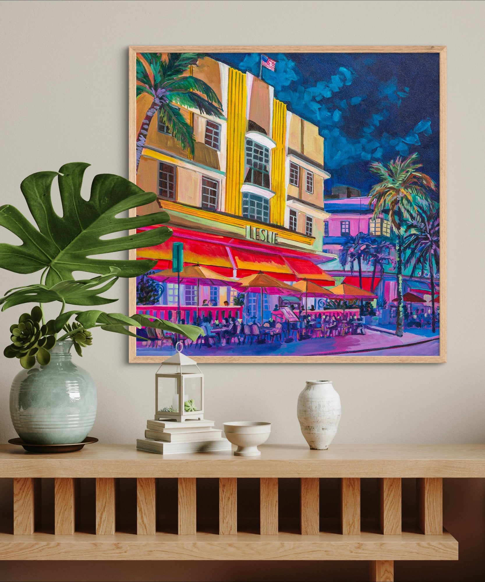 Dramatically lit Leslie hotel in south beach Miami with glowing red awnings is hung on wall above wood shelf with vases and neutral decor.