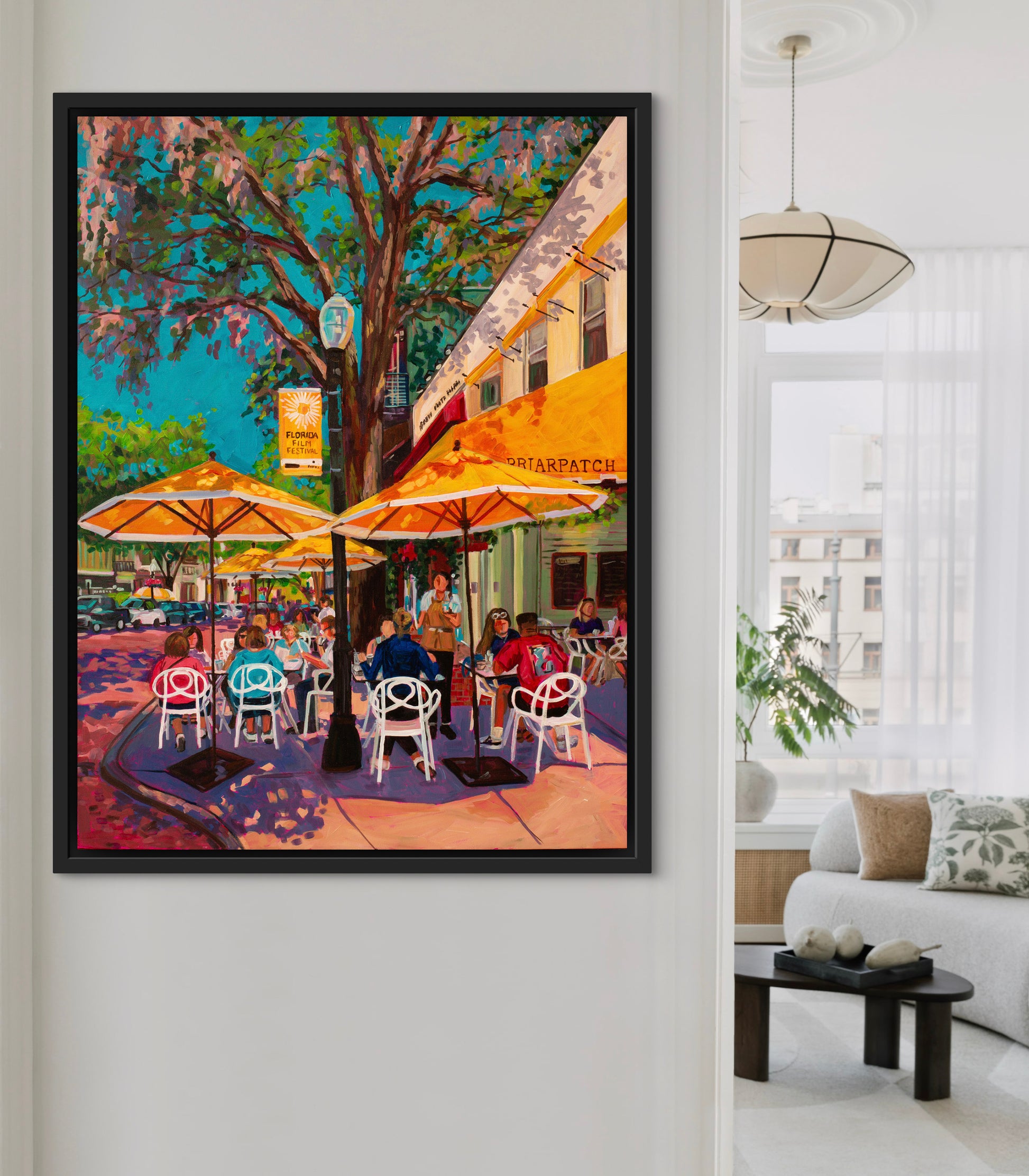Bright white living room area with wall in foreground showing a cheerful outdoor street scene cafe painting with yellow umbrellas.