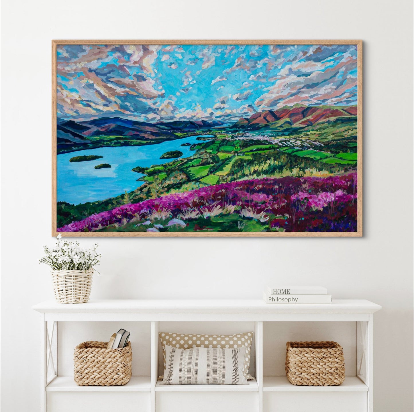 white shelf with baskets, pillows and books, painting on wall above it dreamy landscape with wild heather flowers on cliff overlooking lake with islands, framed by mountains and small town in the middle. 