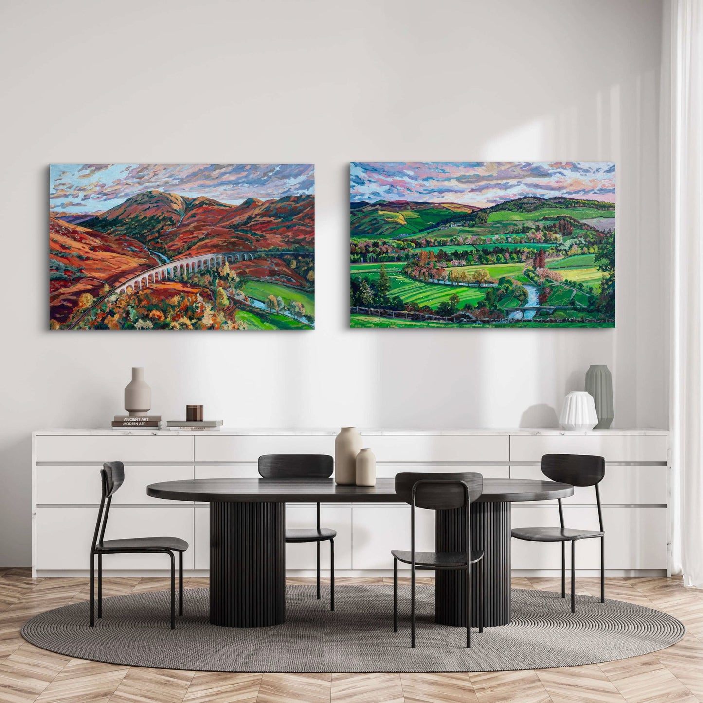 Two dramatic landscape paintings showing rolling hills and in Scotland on wall of minimalist dining room with white credenzas and modern black table and simple chairs.