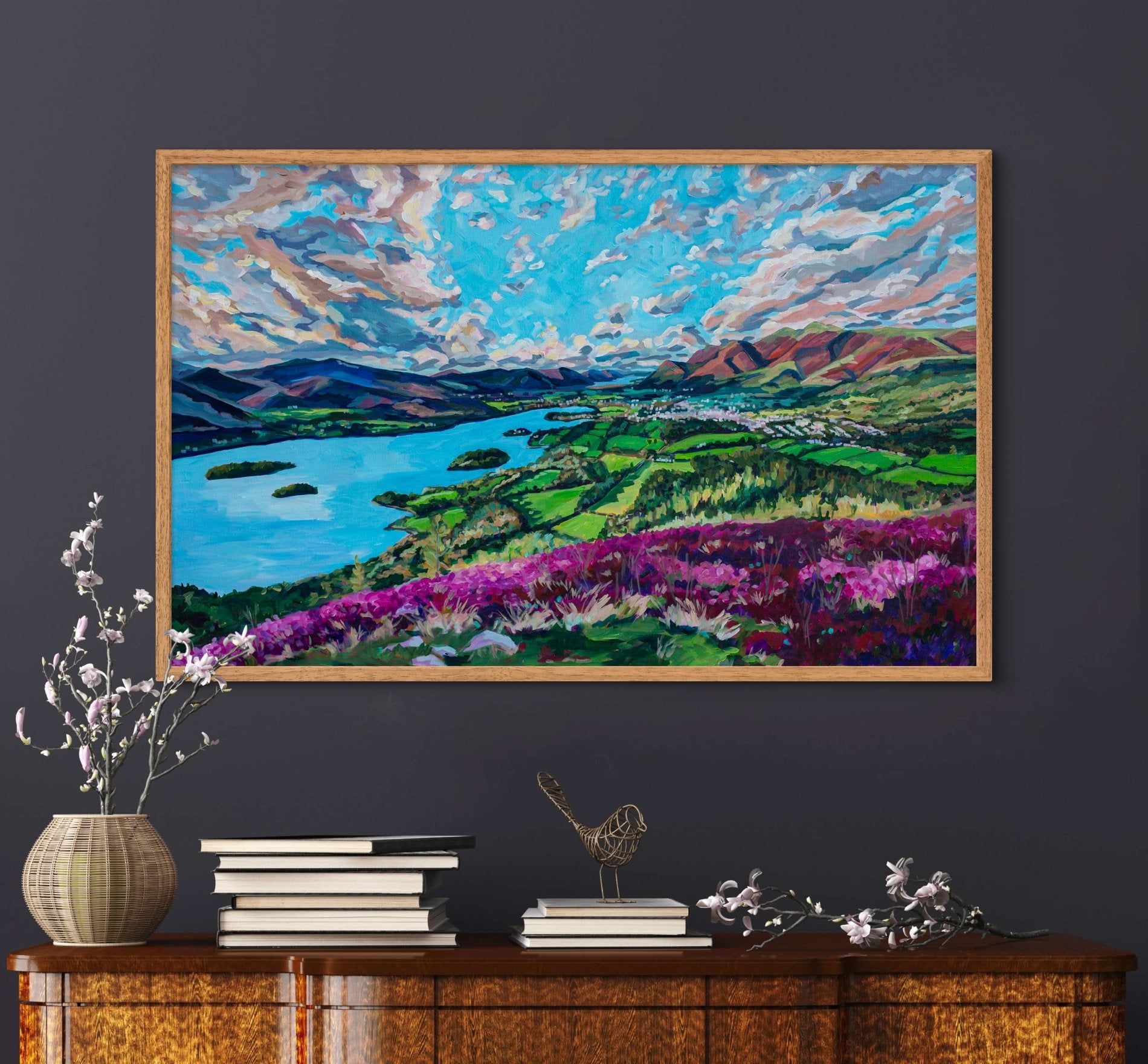 wood credenza with books, and decor in room with dark grey wall and large landscape painting of Derwent Water lake and town of Keswick, Cumbria—UK