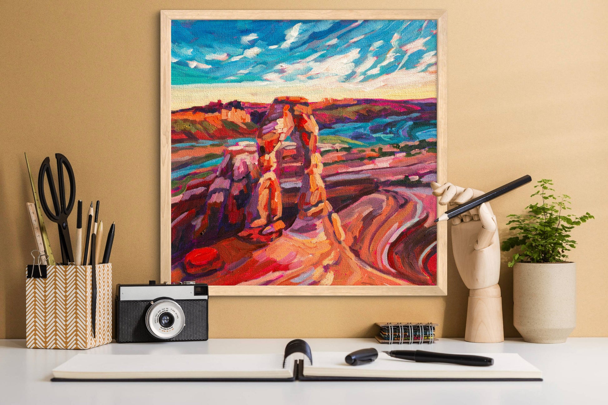 Desk with camera notebook and pencils with a cheerful colorful painting of Delicate arch in Arches National Park.