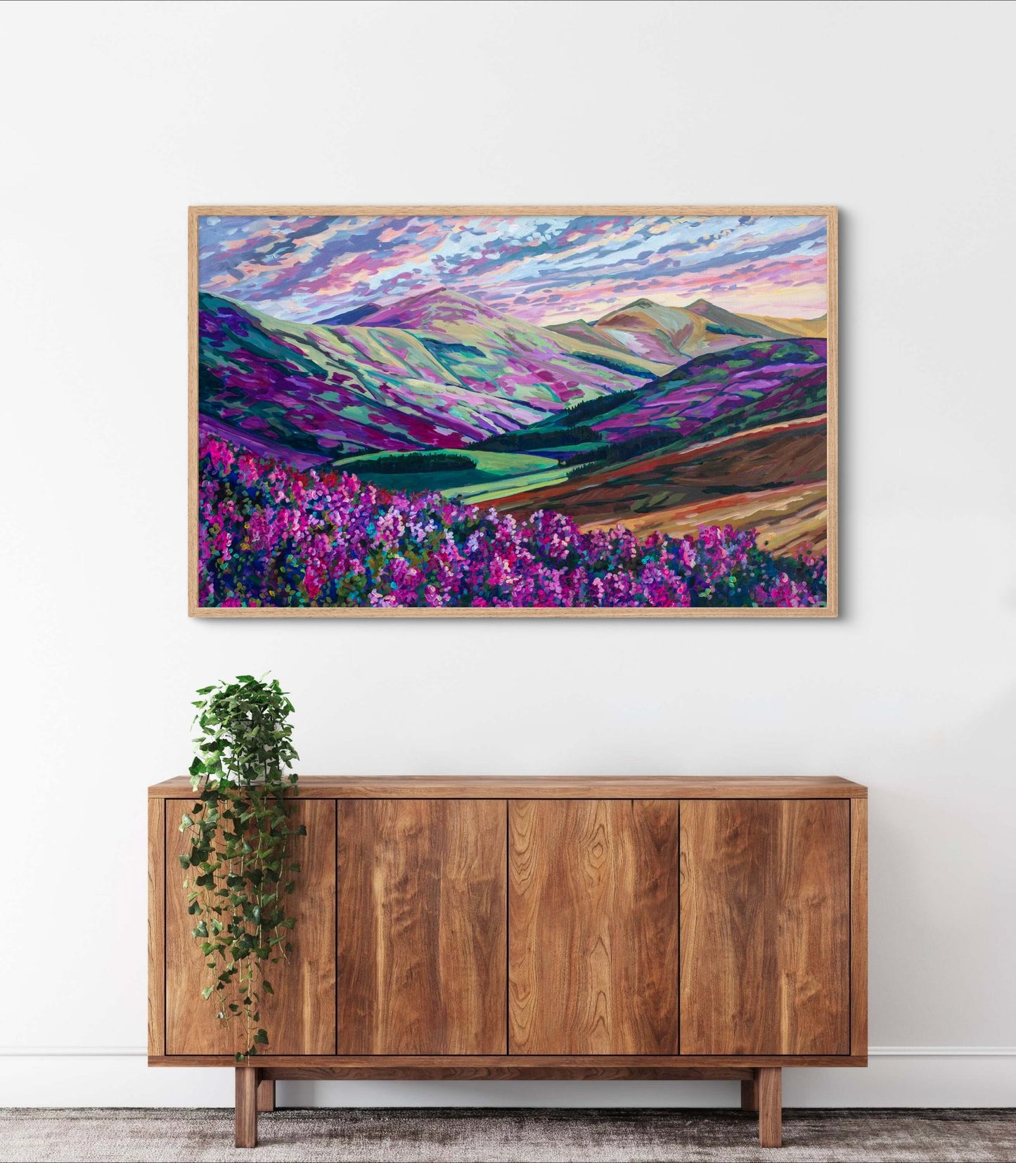 Heather in the Hills, West Linton-Scotland painting, modern vibrant impressionist landscape with purple and pink flowers and mountains over a wood credenza with a potted plant.