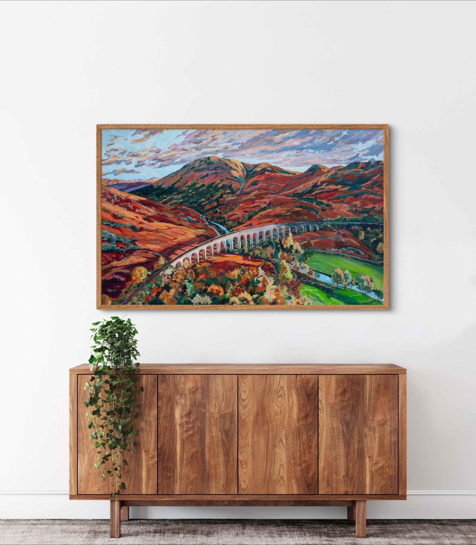 Large statement piece painting showing dramatic lighting near sunrise or sunset with clouds that have some coral and pink colors, above a wood credenza that has a plant on top.