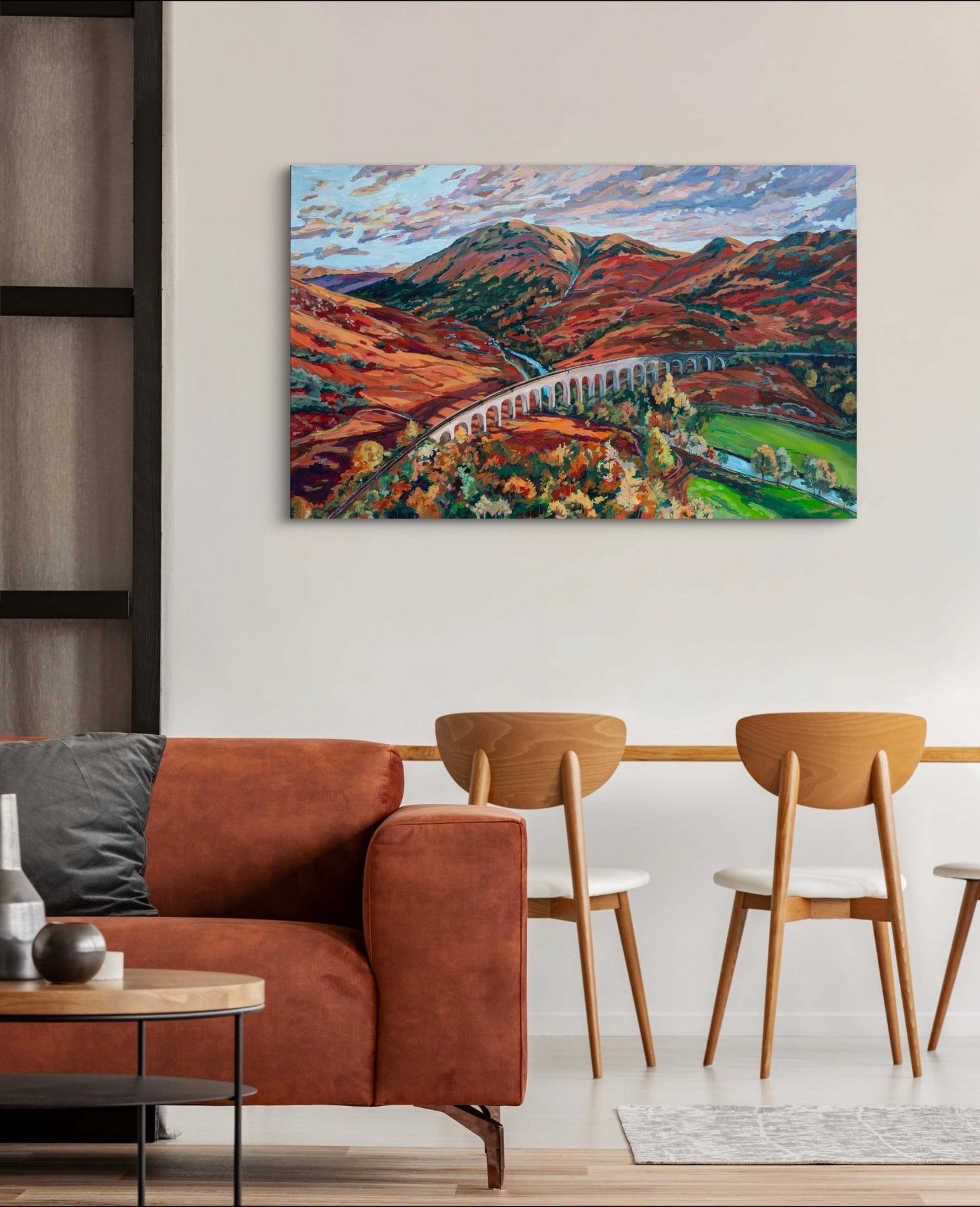 living room area with rust couch, coffee table and dining room table, on wall is a powerful colorful painting matching the coloring showing a rugged landscape of mountains and a stream.