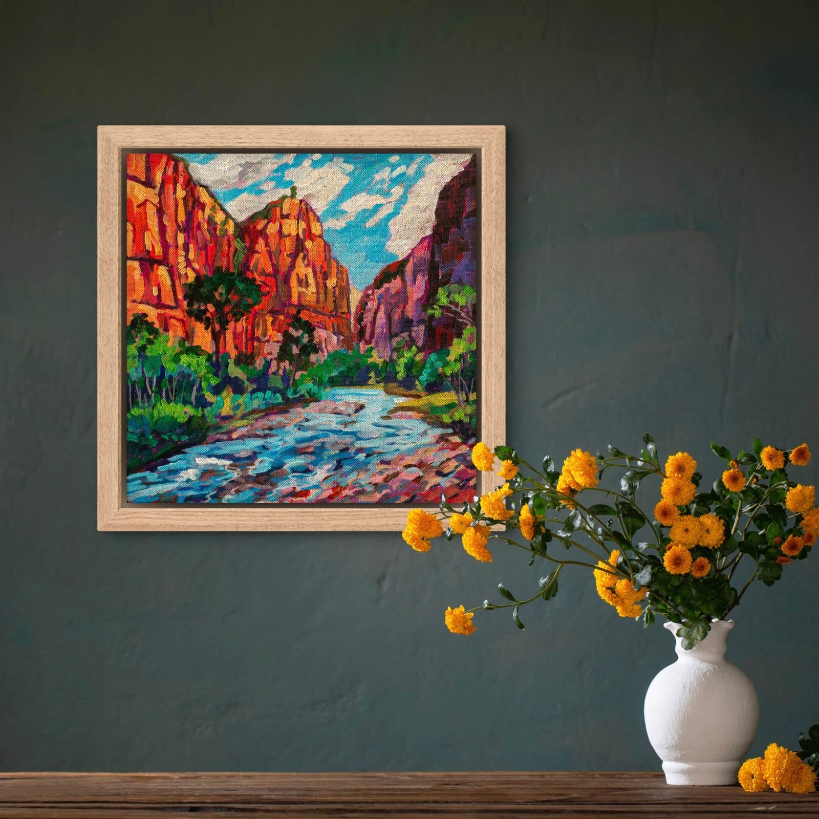 Painting of towering red rock cliff walls above the virgin river in Zion National Park on wall above shelf with vase and orange flowers.