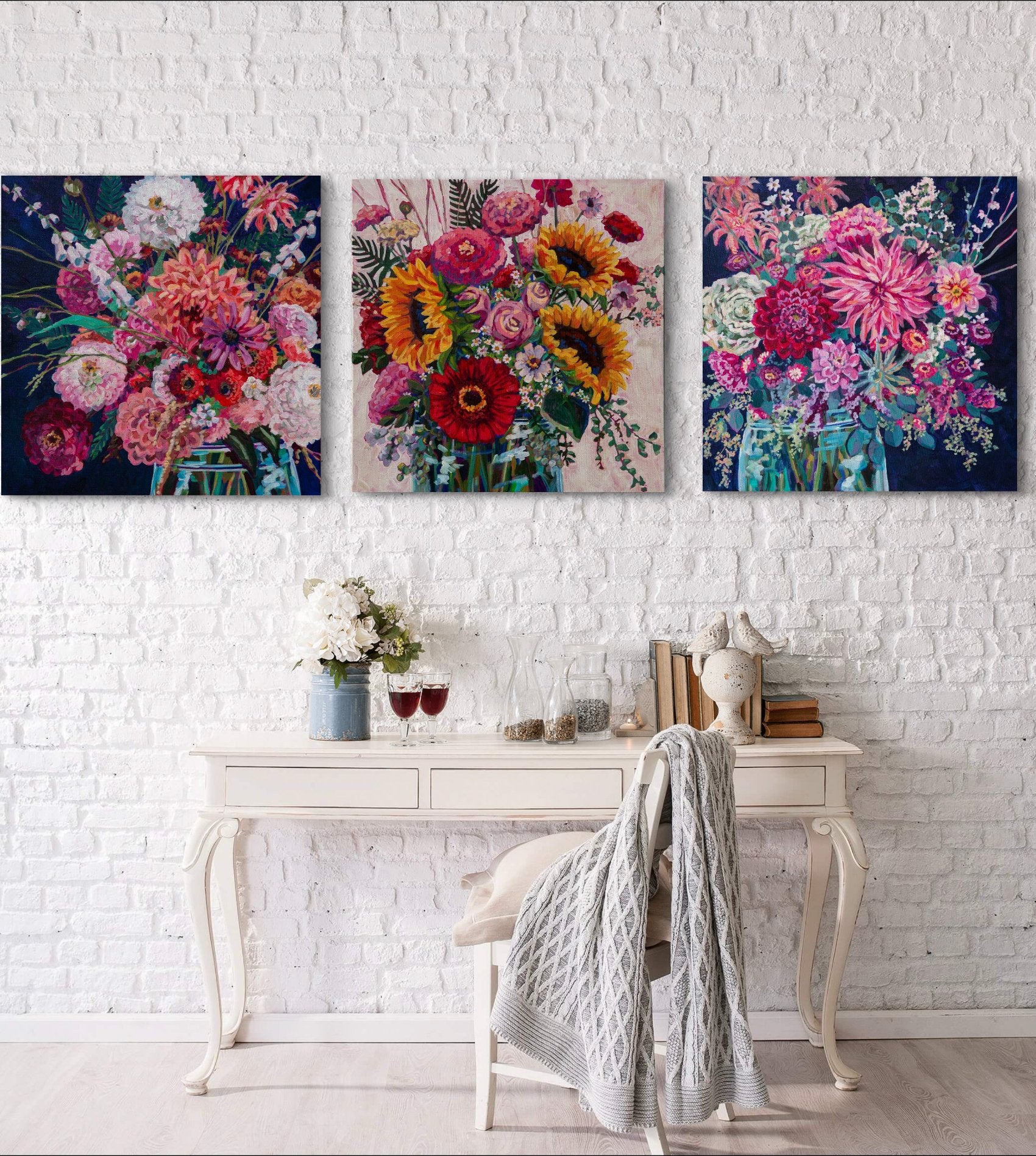 grouping of three vibrant floral arrangement paintings two with navy background, one with cream on a white brick wall in a cozy cottage chic setting with white desk and chair.