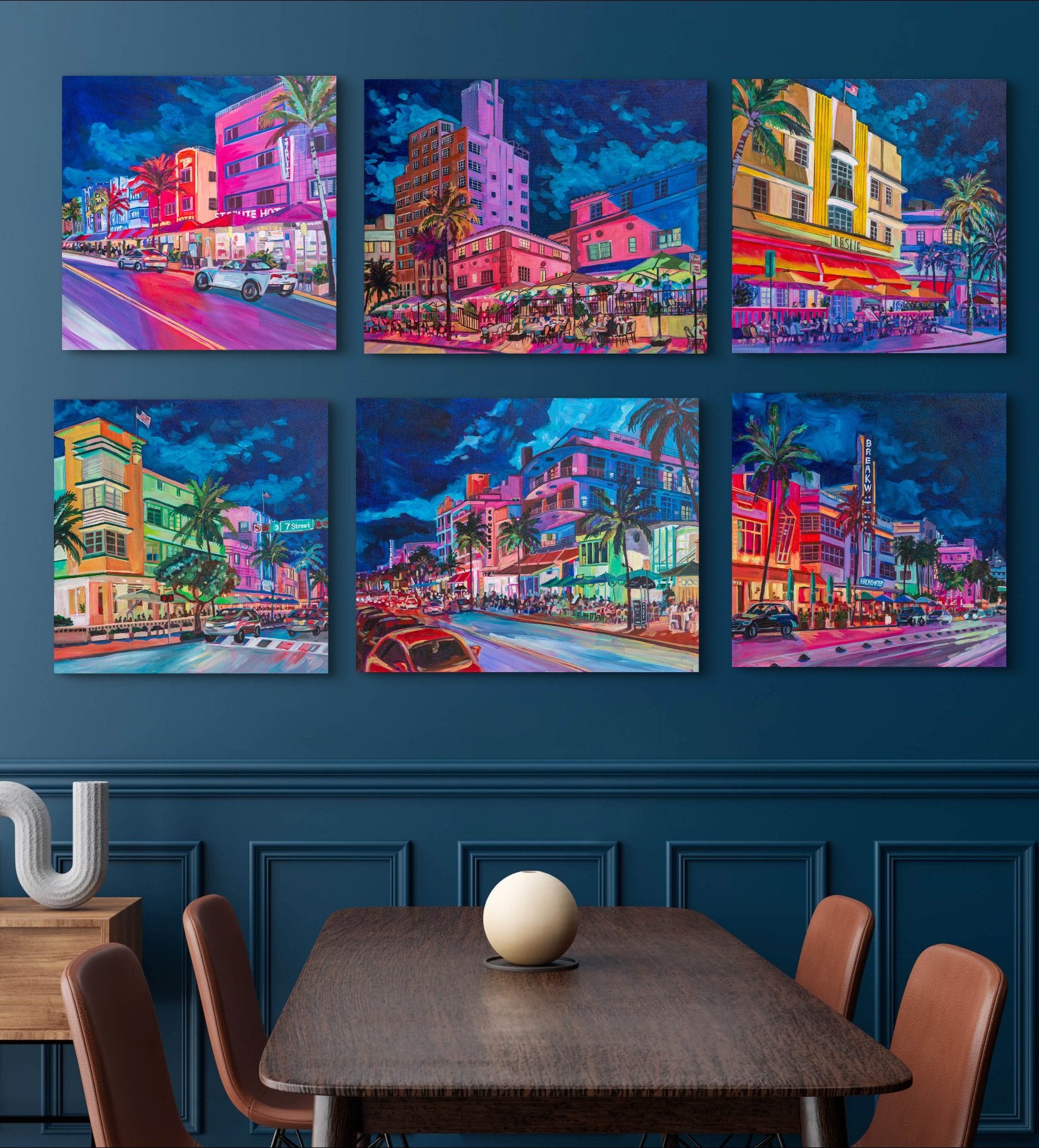 Dramatic dining room with navy walls and a grouping of vibrant paintings of South Beach Miami street scenes with various landmark art deco hotels and restaurants.