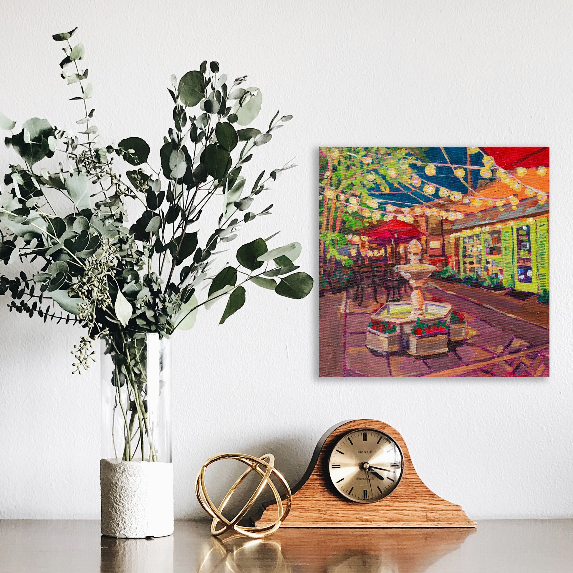 12x12 painting on wall behind dresser with vase and clock: Winter Park Nightlife 2 painting 12x12 vibrant hidden courtyard with strung lights, fountain and tables with umbrellas off Park Ave.